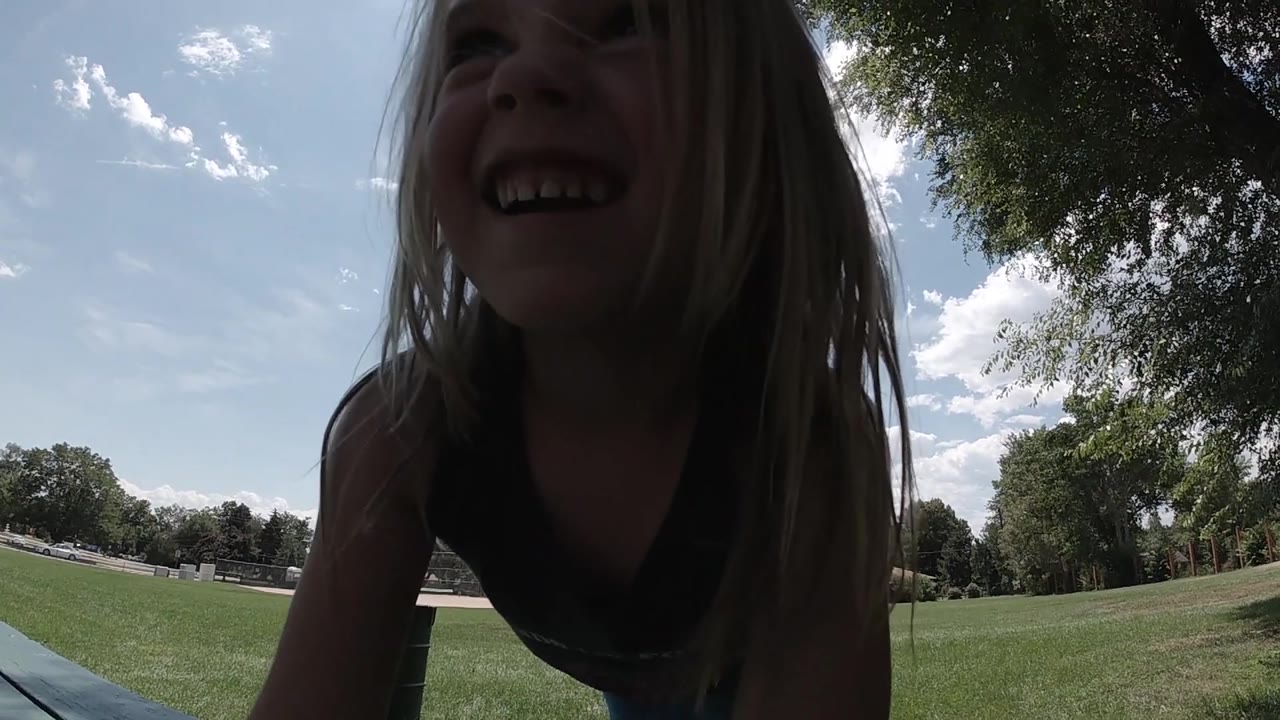 A young girl laughs near the camera, her face shaded by backlight while the Colorado park lawn and trees stay bright behind her.