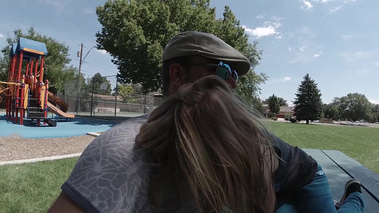 At the start of the Colorado clip, a father in sunglasses holds close to a blonde girl beside a green picnic table with colorful playground equipment behind them.