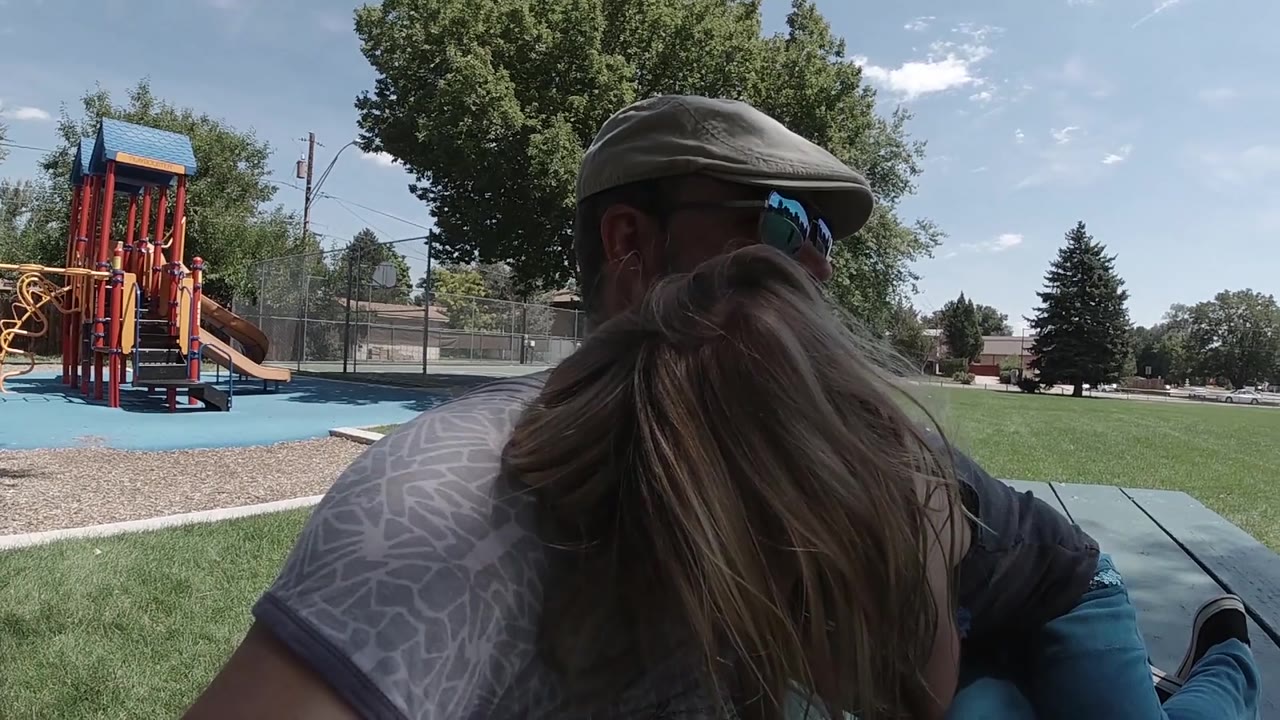 The father and blonde girl remain close in the foreground, with orange playground posts, green grass, and bright sky visible across the Colorado park.