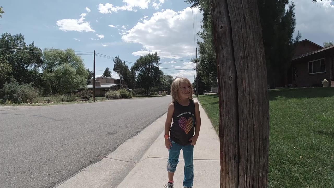 The girl remains on the sidewalk near the right-side tree trunk, looking off to her left while the road and low homes stretch behind her in Colorado.
