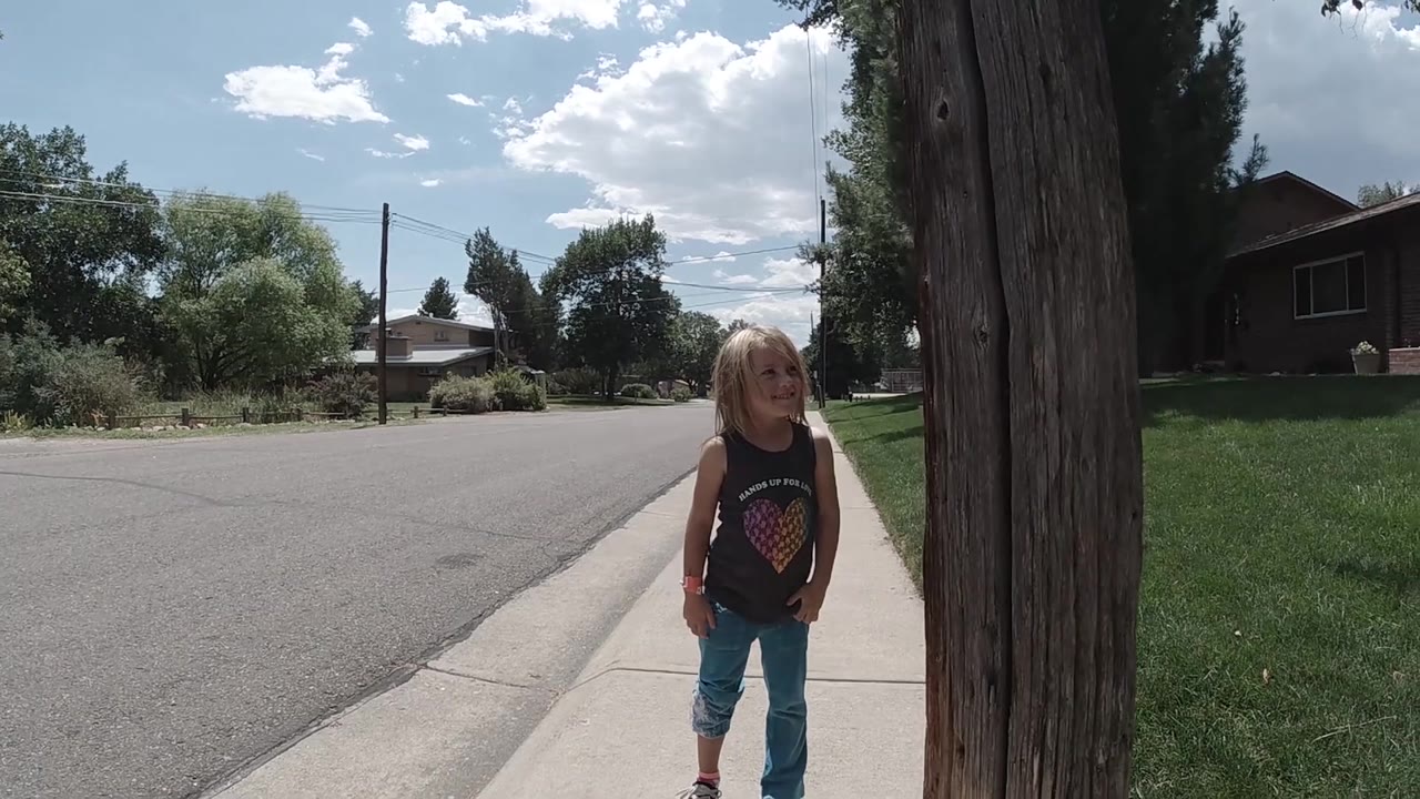 A young girl stands centered on the sidewalk with the tree trunk close at right, the residential road at left, and sunlit Colorado greenery in the background.