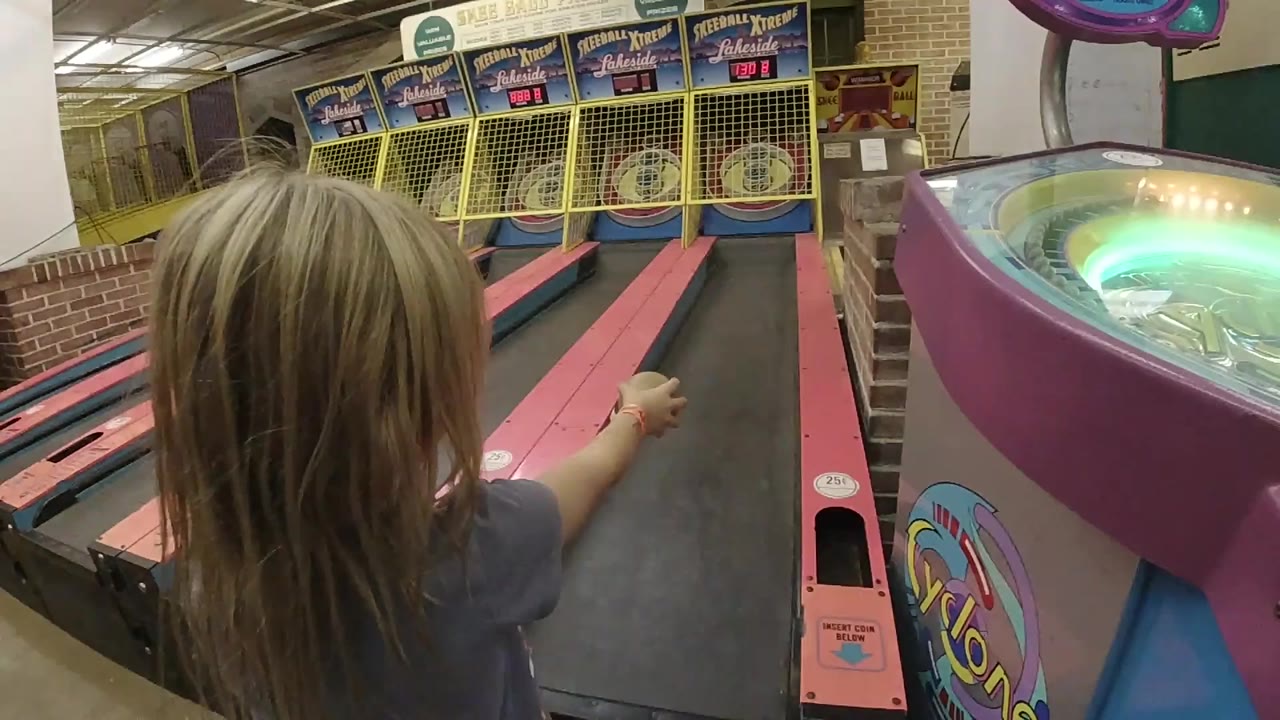 At the start of the Colorado arcade clip, a blonde young girl points toward the skee-ball targets from the near end of a pink lane.