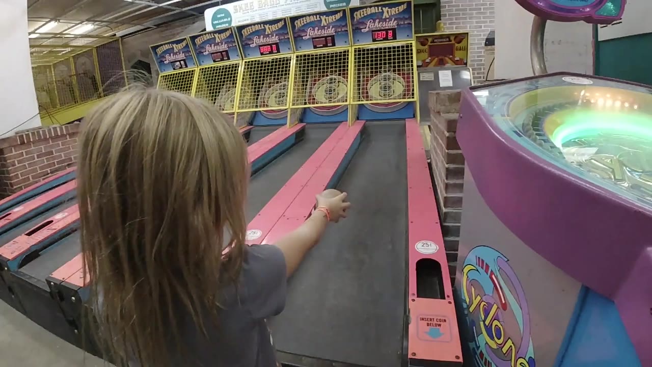The girl remains near the lane edge with her hand extended, facing the skee-ball targets in the Colorado indoor arcade.