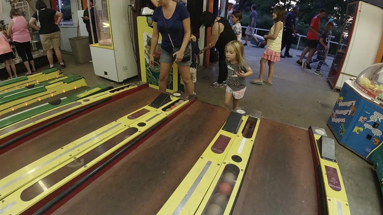At the start of the clip, a young girl stands near the front of a yellow skee-ball lane in a Colorado arcade while an adult woman and other visitors are visible around the machines.