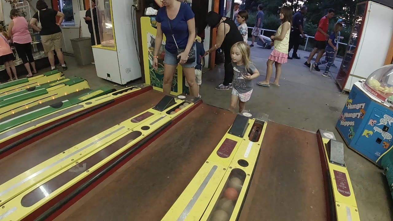 The girl remains beside the yellow skee-ball lane while the adult woman leans nearby and arcade visitors continue moving in the background.