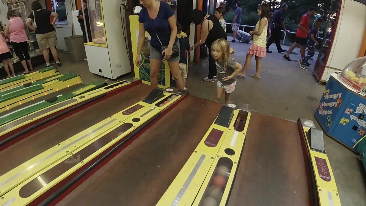The child faces the skee-ball area with the woman close behind her, while the arcade floor and doorway remain busy with visible people.