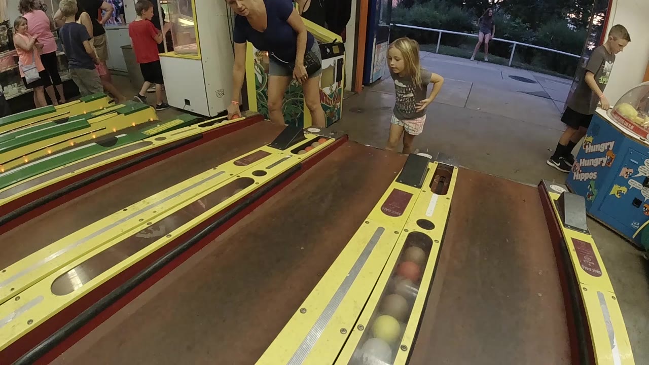 The girl faces the skee-ball lanes while an adult woman stands close by and other arcade visitors remain near the machines in the background.