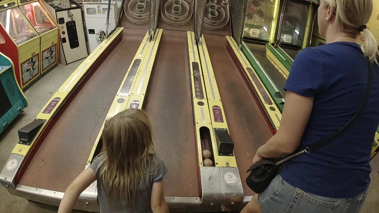 In a Colorado arcade, a mother in a blue shirt stands beside a young blonde daughter facing two skee-ball lanes at the start of the play moment.