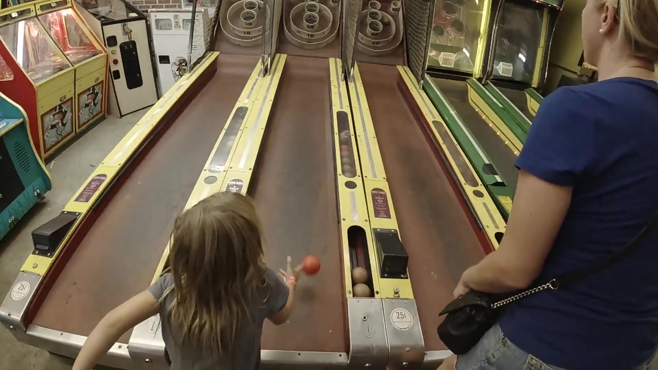 A red skee-ball rolls up the right lane as the child and mother remain in the foreground of the indoor Colorado arcade scene.