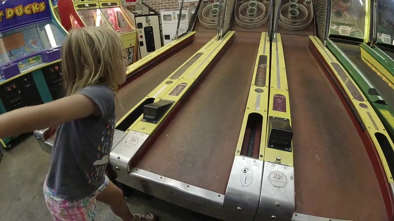 A young blonde girl in Colorado stands at the left edge of a skee-ball lane with her arm extended near the ball return and arcade machines around her.