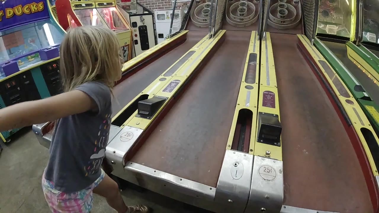 The child remains near the left side of the skee-ball ramp in Colorado, reaching across the foreground while the long arcade lane leads toward target rings.