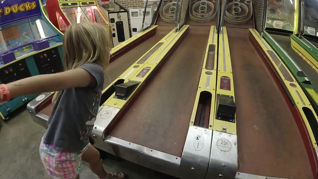 A blonde child in Colorado leans toward the skee-ball machine with her arm out, while neighboring arcade games and bright lane borders fill the background.
