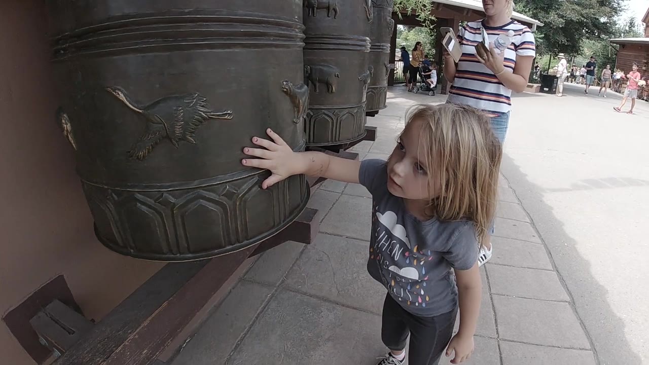 The Denver frame holds close on the girl's outstretched hand beside a raised animal figure on the dark metal display, with visitors faintly visible down the walkway.