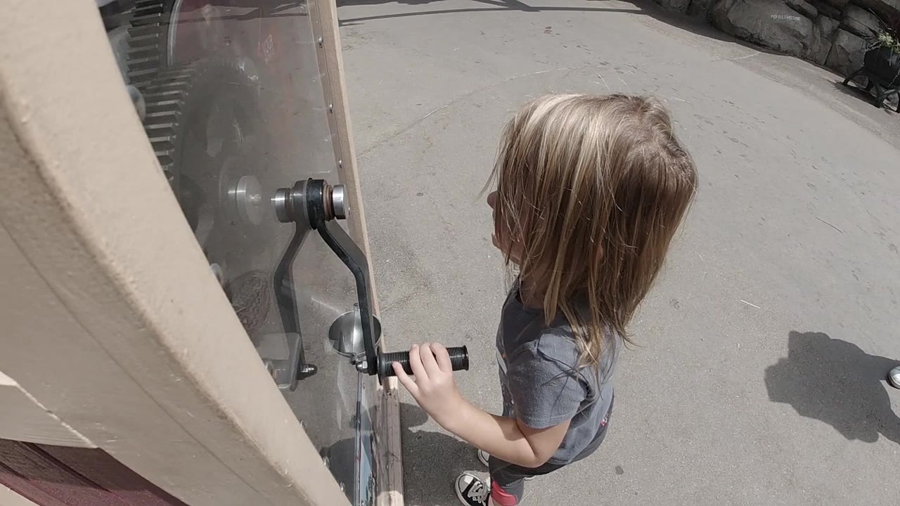 Blonde child holding a black crank handle beside a clear panel.