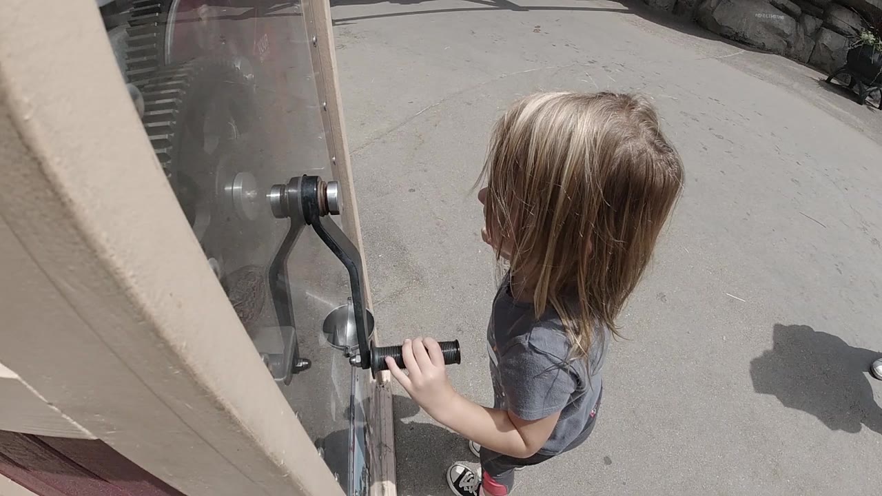 The child remains beside the clear machine panel in Denver, with one hand on the black handle and bright sun casting shadows on the pavement.