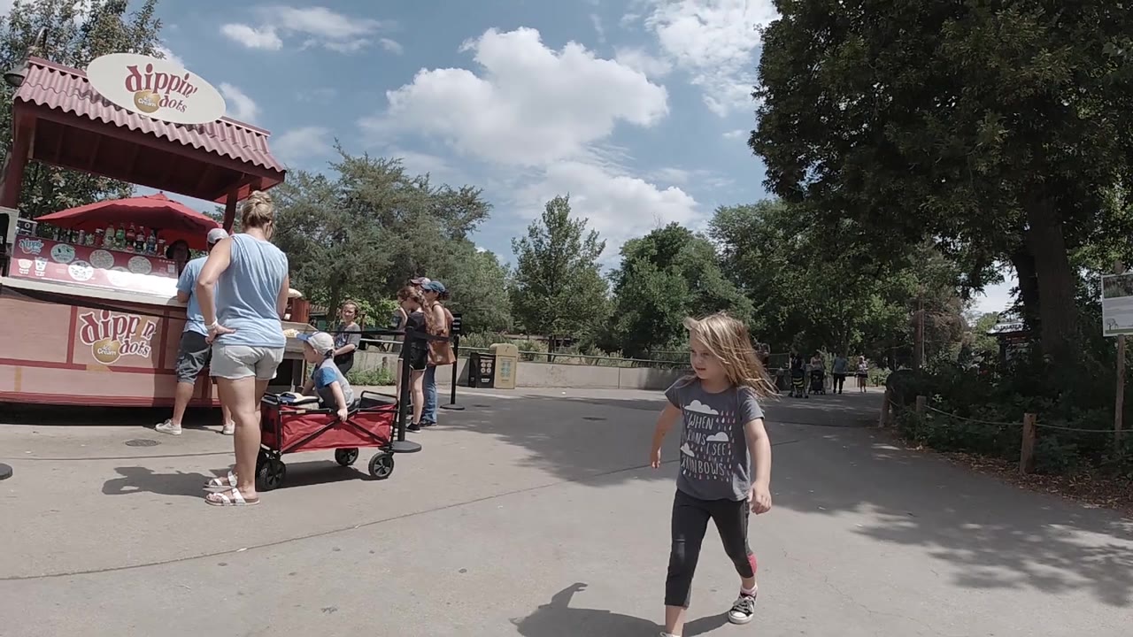 At Denver Zoo, the child nears the right edge of the frame while the Dippin' Dots stand, a stroller or wagon area, adults, trees, and bright clouded sky stay in view.