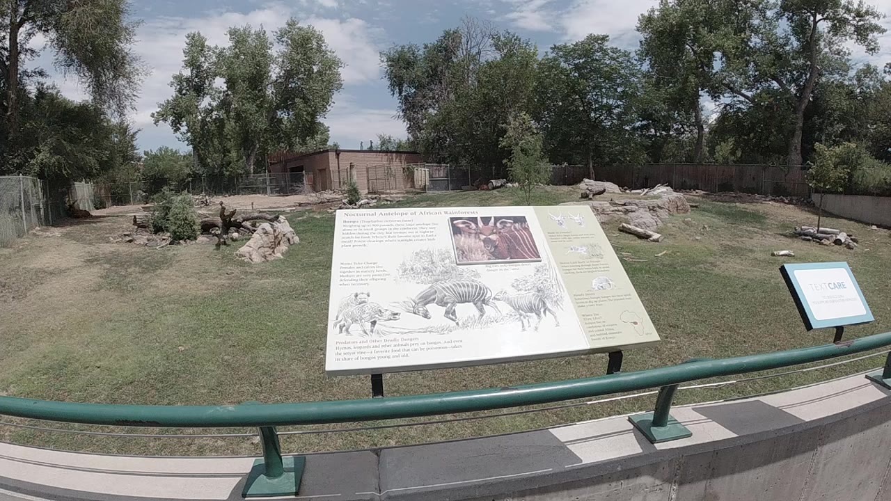 Zoo interpretive sign, green rail, grass, rocks, trees, and blue sky.