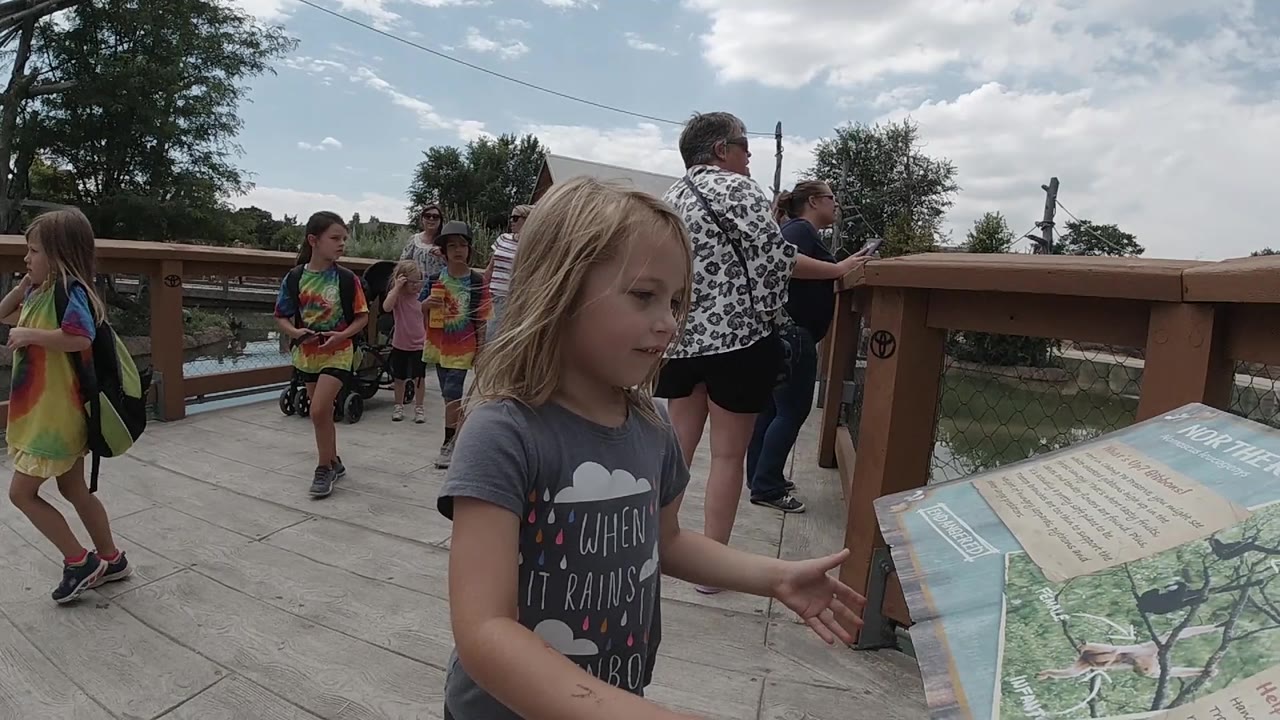 The girl faces the sign with one hand near the board as visitors and backpacks appear behind the wooden railing area in Colorado.
