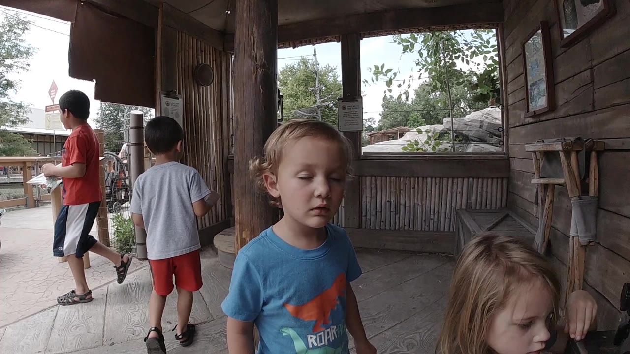 At a Colorado zoo, a blond boy in a blue shirt stands near the camera while a young girl is partly visible beside a wooden wall and two boys look out from the walkway.