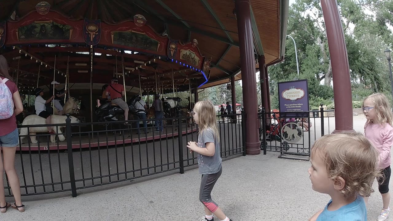 A blonde child in gray stands closer to the fence as another child in blue is in the foreground and the carousel pavilion fills the left side of the Colorado park frame.