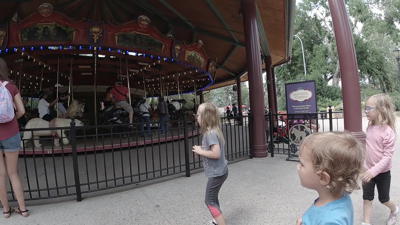 Three children are visible on the paved path beside the black fence, with carousel horses, seated riders, blue lights, and trees behind the covered ride.