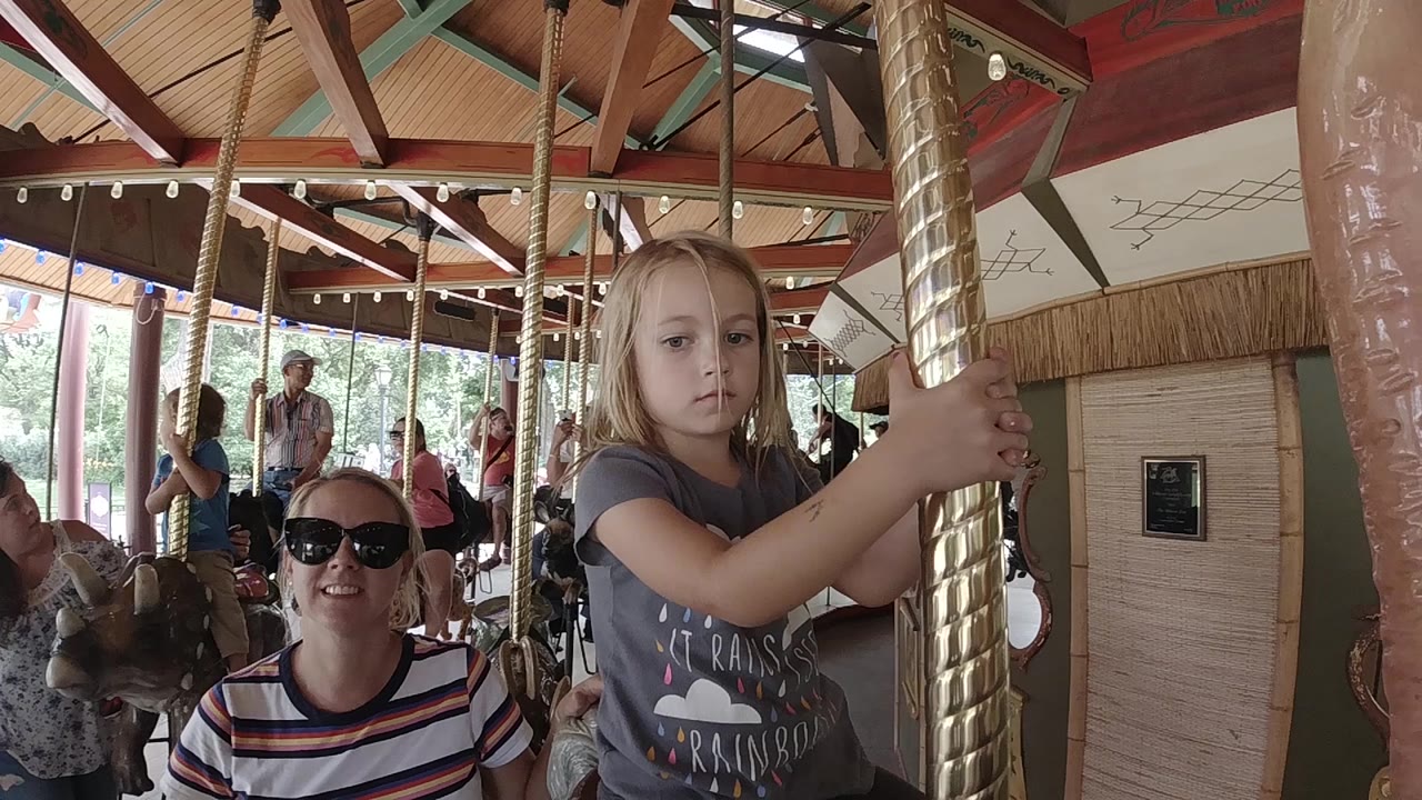 One second in, the girl remains close to the gold carousel pole while the adult woman faces the camera from the lower left and riders appear behind them.