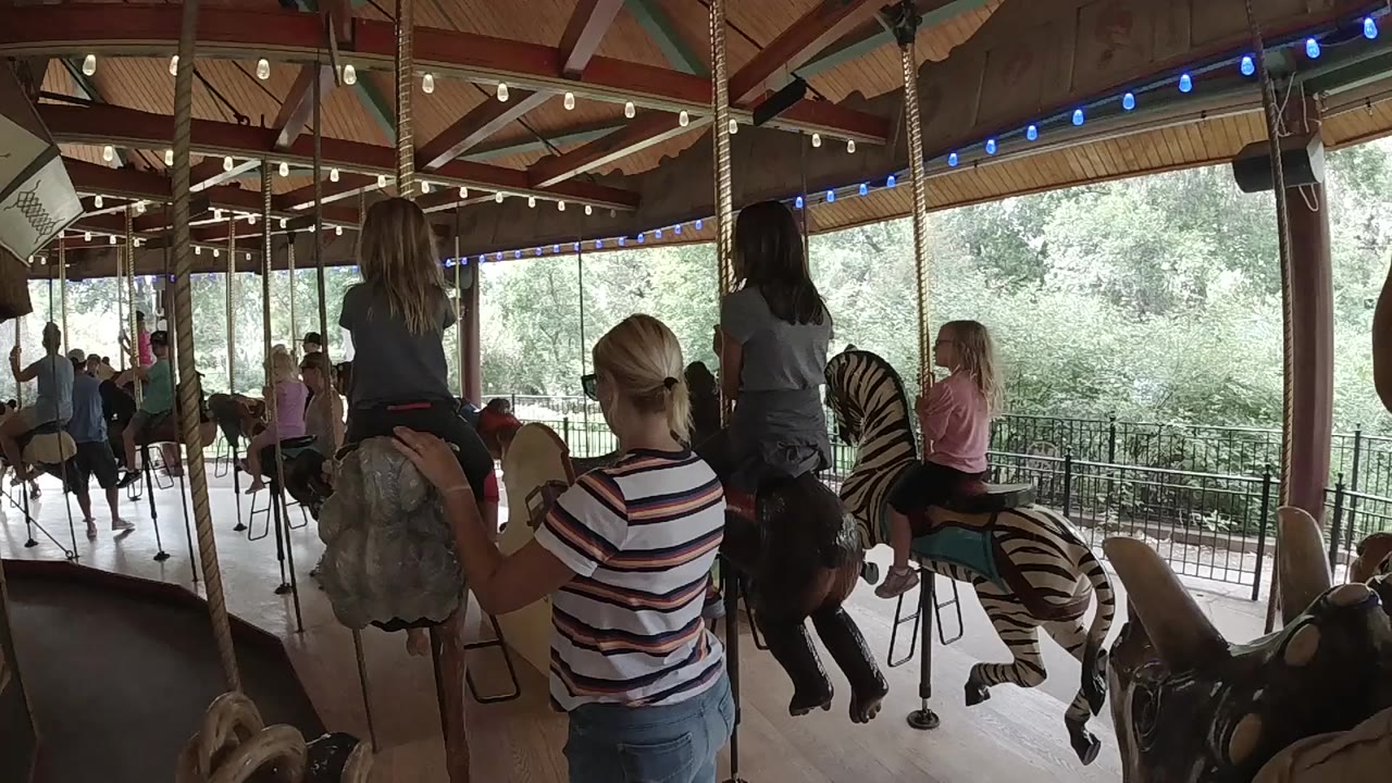 Opening frame of a covered carousel in Colorado with children on painted animals, adults nearby, blue bulbs overhead, and trees beyond the railings.