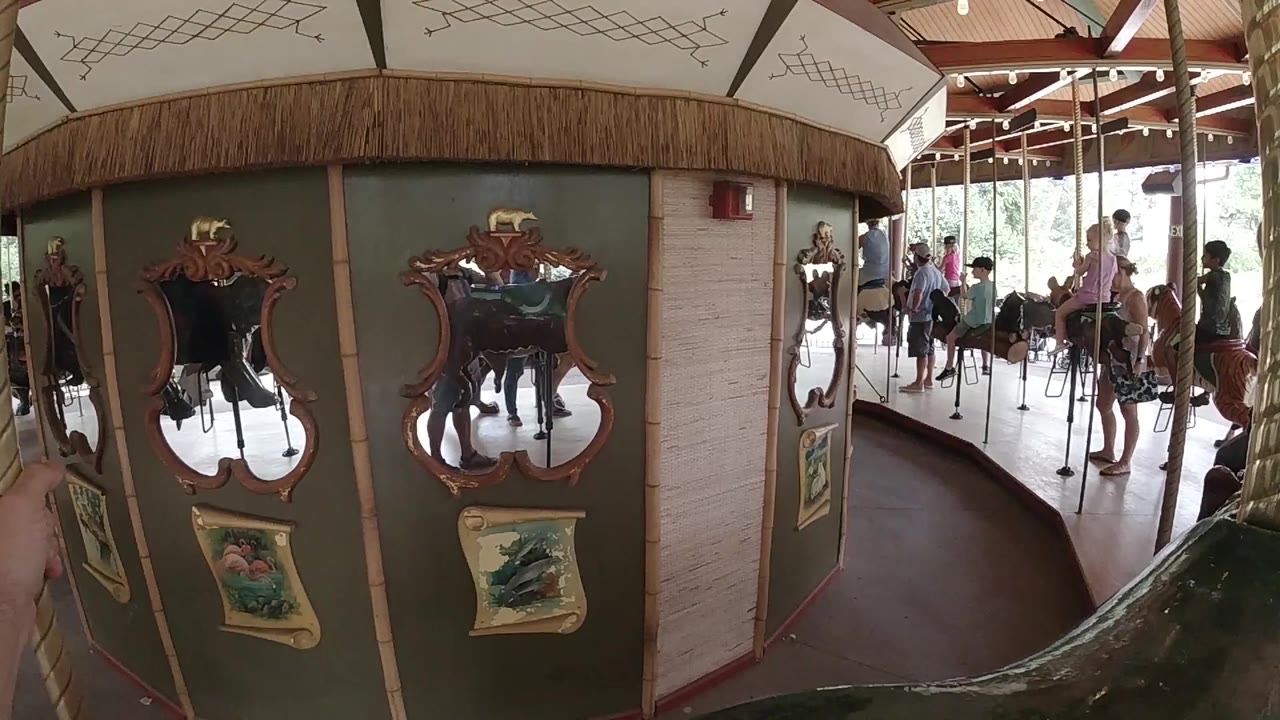 At 0:00, ornate carousel mirrors and painted panels fill the foreground in Colorado, with people and riders visible beyond the curved interior wall.
