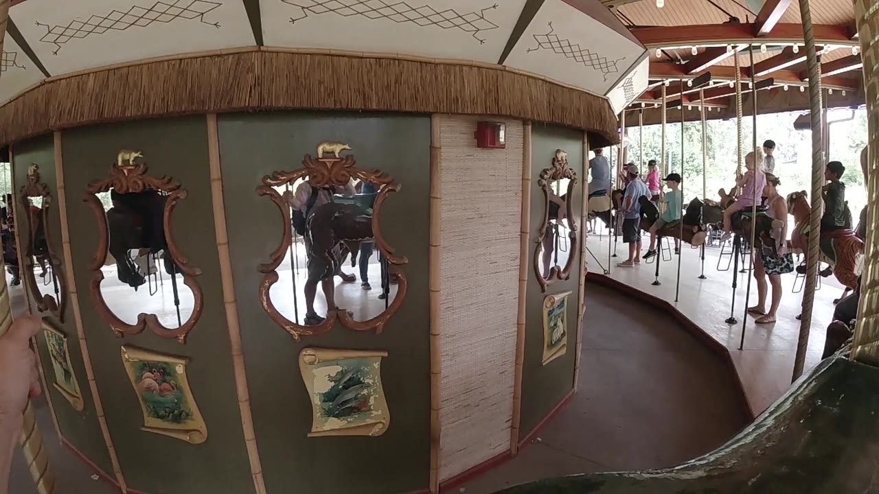 At 0:02, two ornate mirrors on the carousel center structure reflect the ride area while people stand and ride in the bright background.