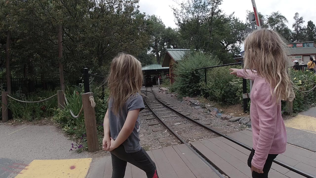 Two young girls in gray and pink tops stand near narrow train tracks and a wooden crossing path in Colorado, United States.