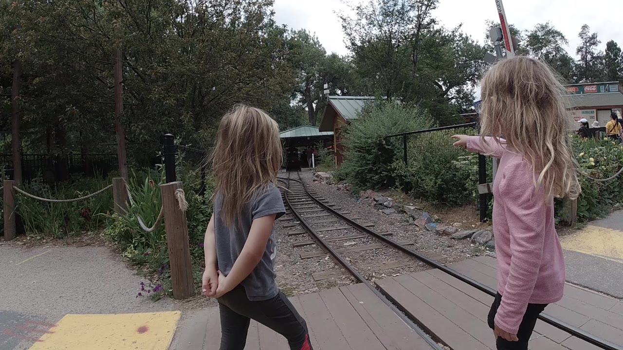 The girl in a pink long-sleeve shirt points across the narrow tracks while the other child faces the zoo railway area in Colorado, United States.