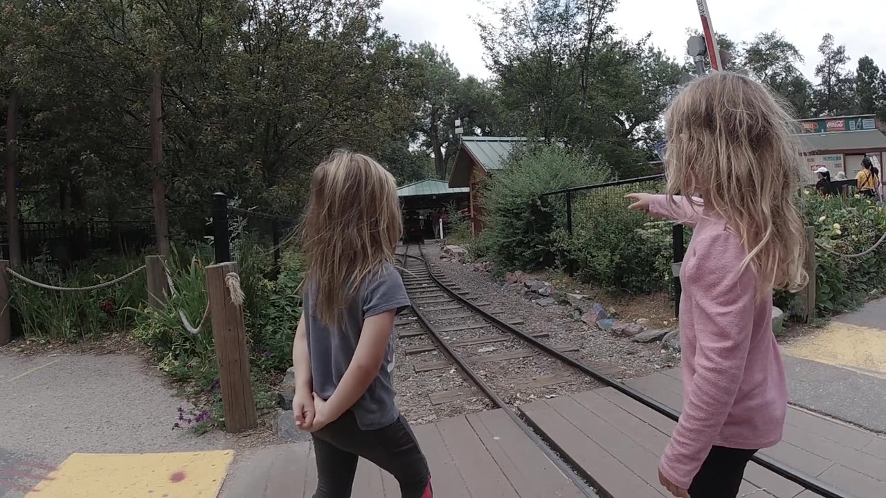 Two children stand at the edge of a path and look down the narrow zoo train tracks, with shrubs and a low building behind them in Colorado, United States.