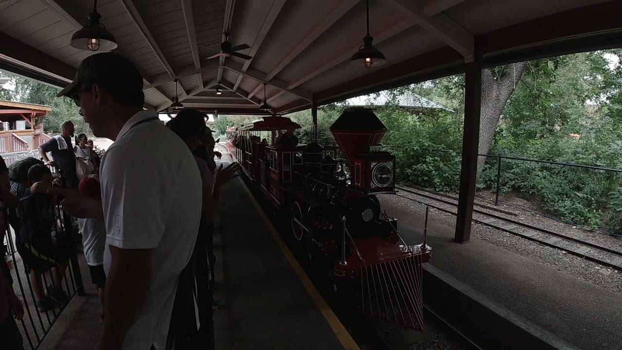 At a Colorado zoo platform, a red train approaches under a roof while adults wait at the left and green trees line the track.