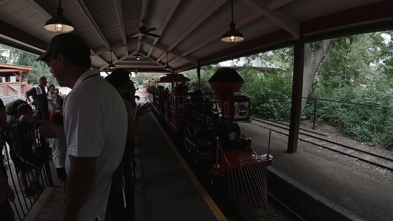 The red train moves nearer along the platform, with a man in a white shirt in the foreground and passengers gathered under the shaded roof.