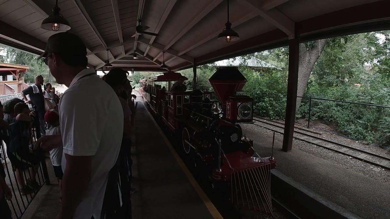 A red locomotive front reaches the platform area in Colorado, with adults visible on the left and dense green foliage beyond the tracks.
