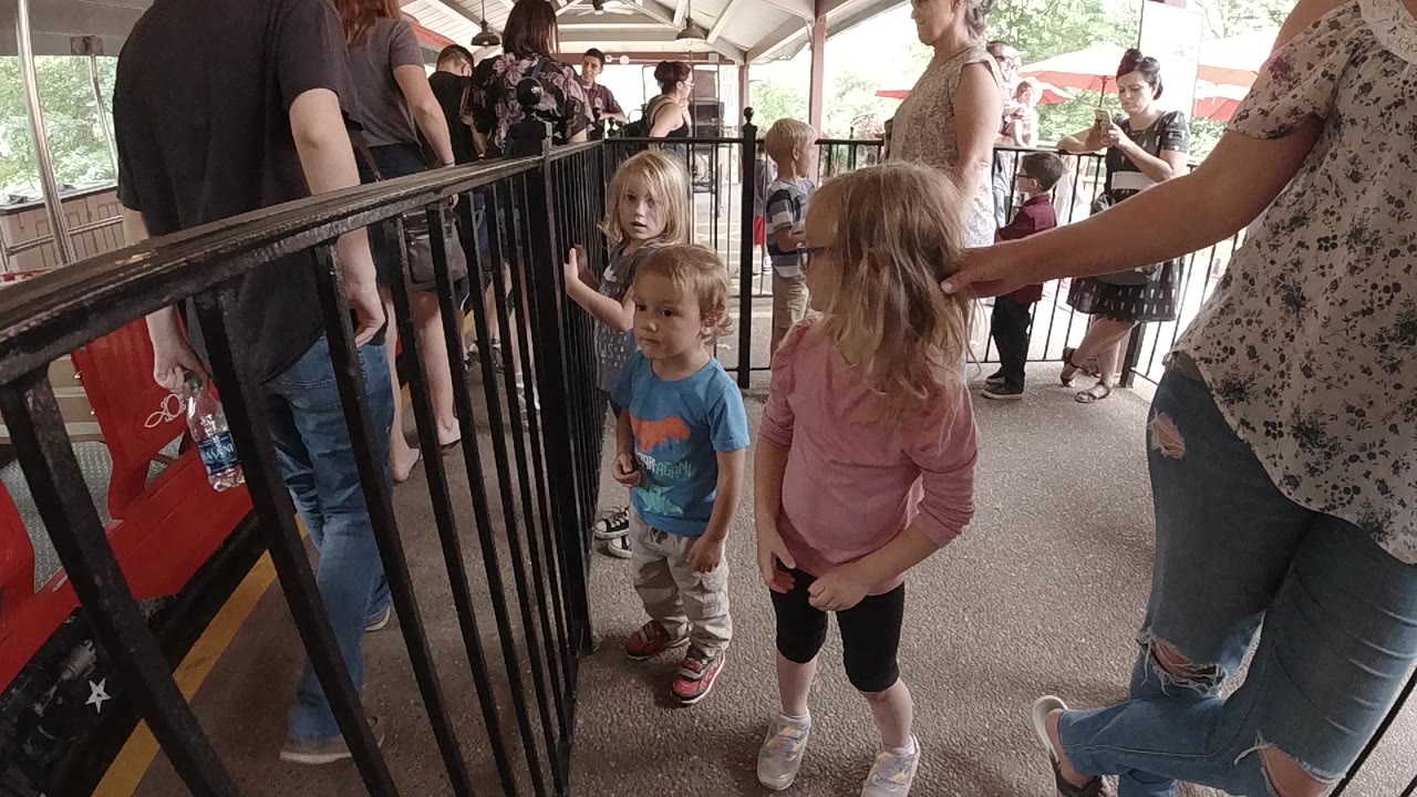 At the start of the Colorado clip, two young children stand beside black metal railings in a covered zoo train queue while adults wait around them.