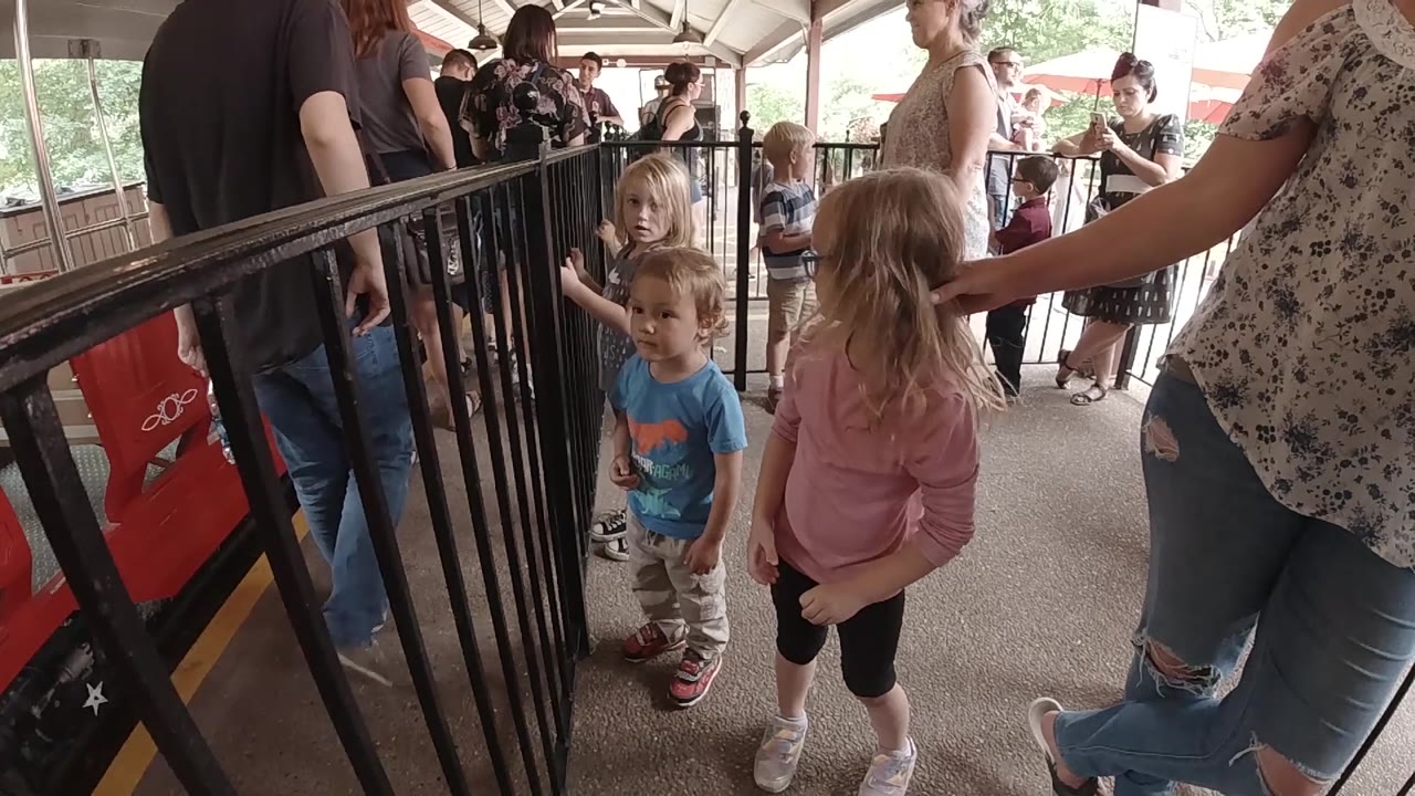 The girl in a pink shirt and the boy in a blue shirt wait beside black railings, with a red train car edge and clustered adults visible under the platform roof.