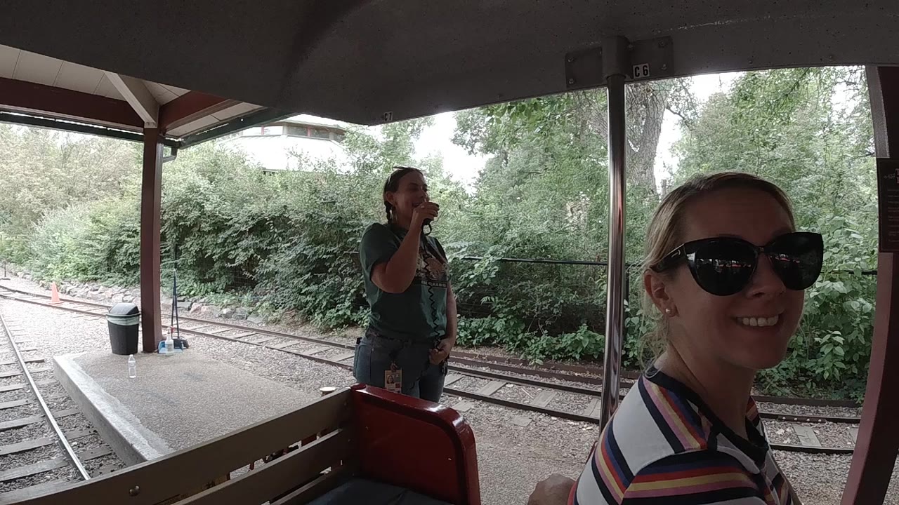 At 0:01 in Colorado, the seated woman remains close to the right side of frame, smiling beside the train car opening as rails and leafy bushes run behind the platform.