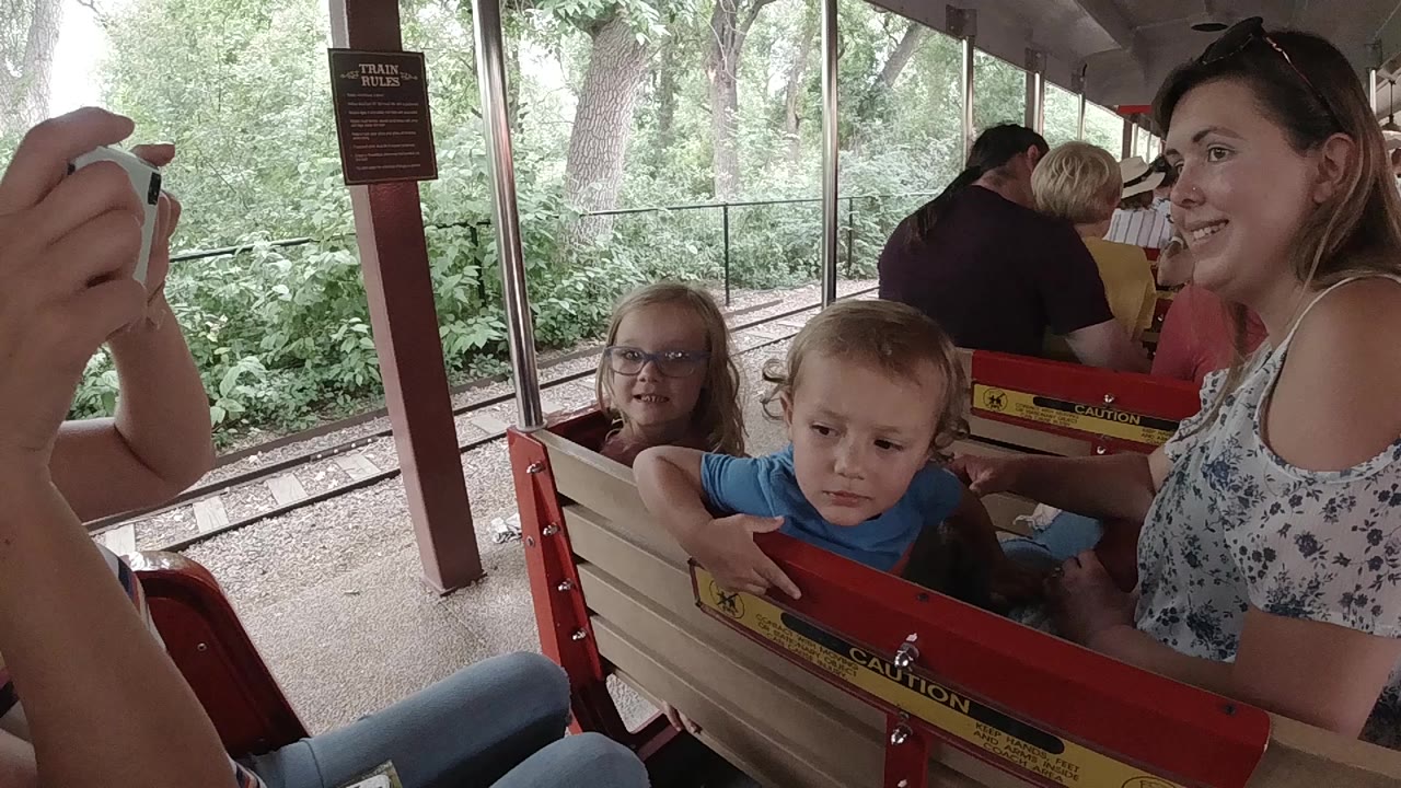 In Colorado, a woman sits beside a young boy and girl on a red and cream covered ride bench with trees and track visible outside.
