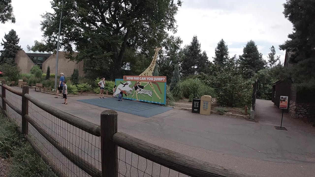 Wide Colorado zoo scene with visitors near the left side of the path and children around the blue mat beside a colorful animal-themed jumping sign.