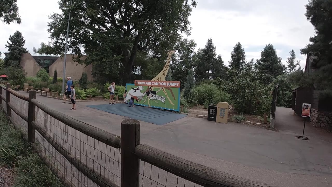 Colorado zoo walkway framed by a foreground fence, with a blue mat, colorful jumping challenge sign, trees, shrubs, bins, and people visible near the play area.