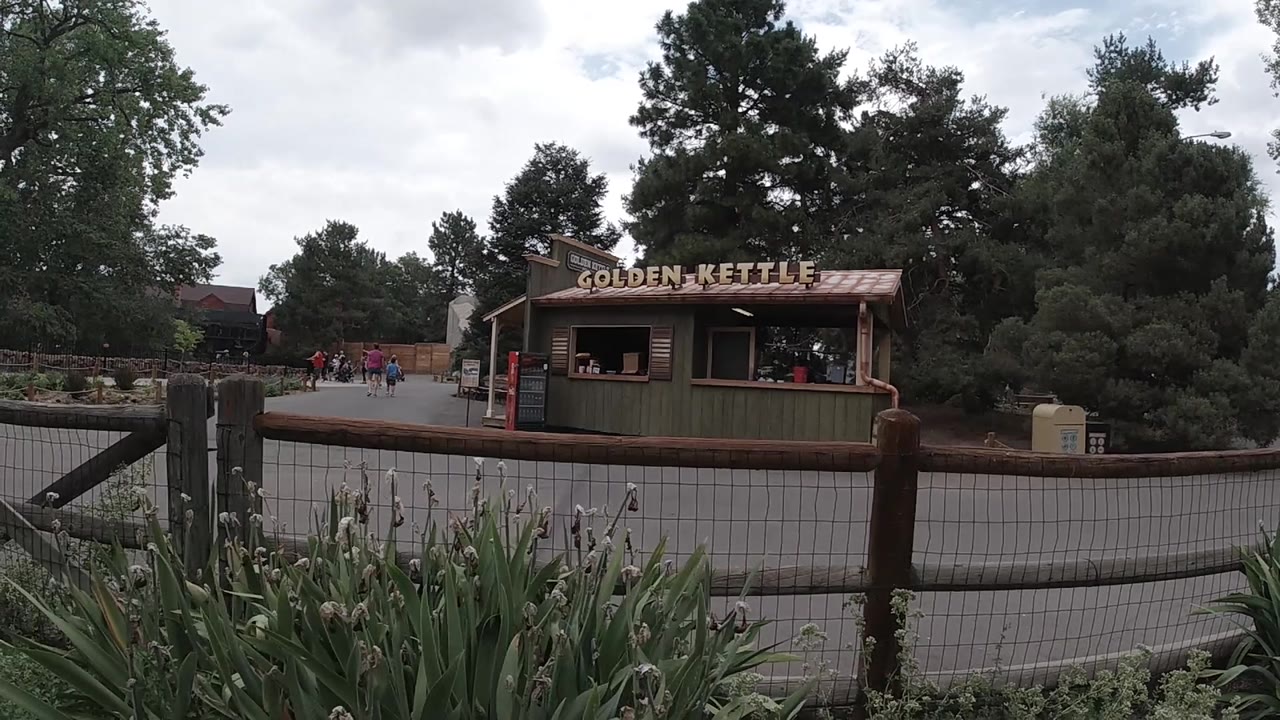 A wide Colorado zoo frame shows the Golden Kettle sign above the green kiosk, with the wooden fence crossing the foreground and visitors small on the left path.