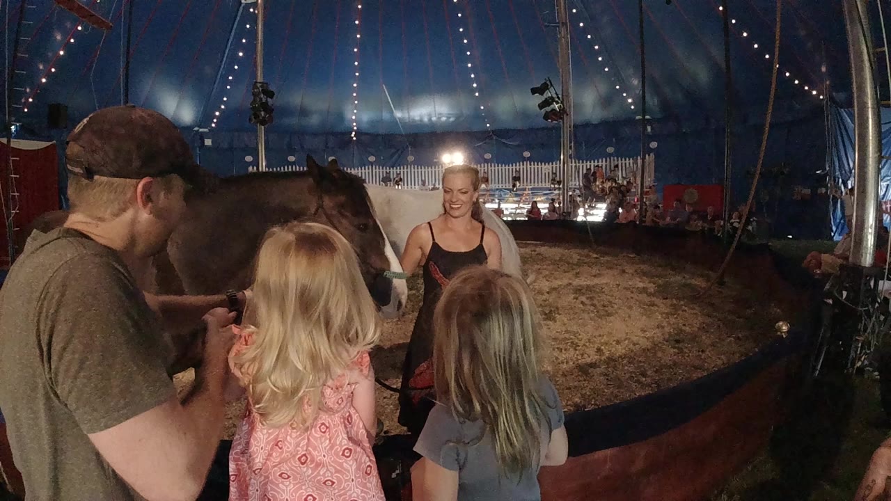 In Colorado, a man and two girls face a woman near a dark horse inside a blue circus tent with arena lights behind them.