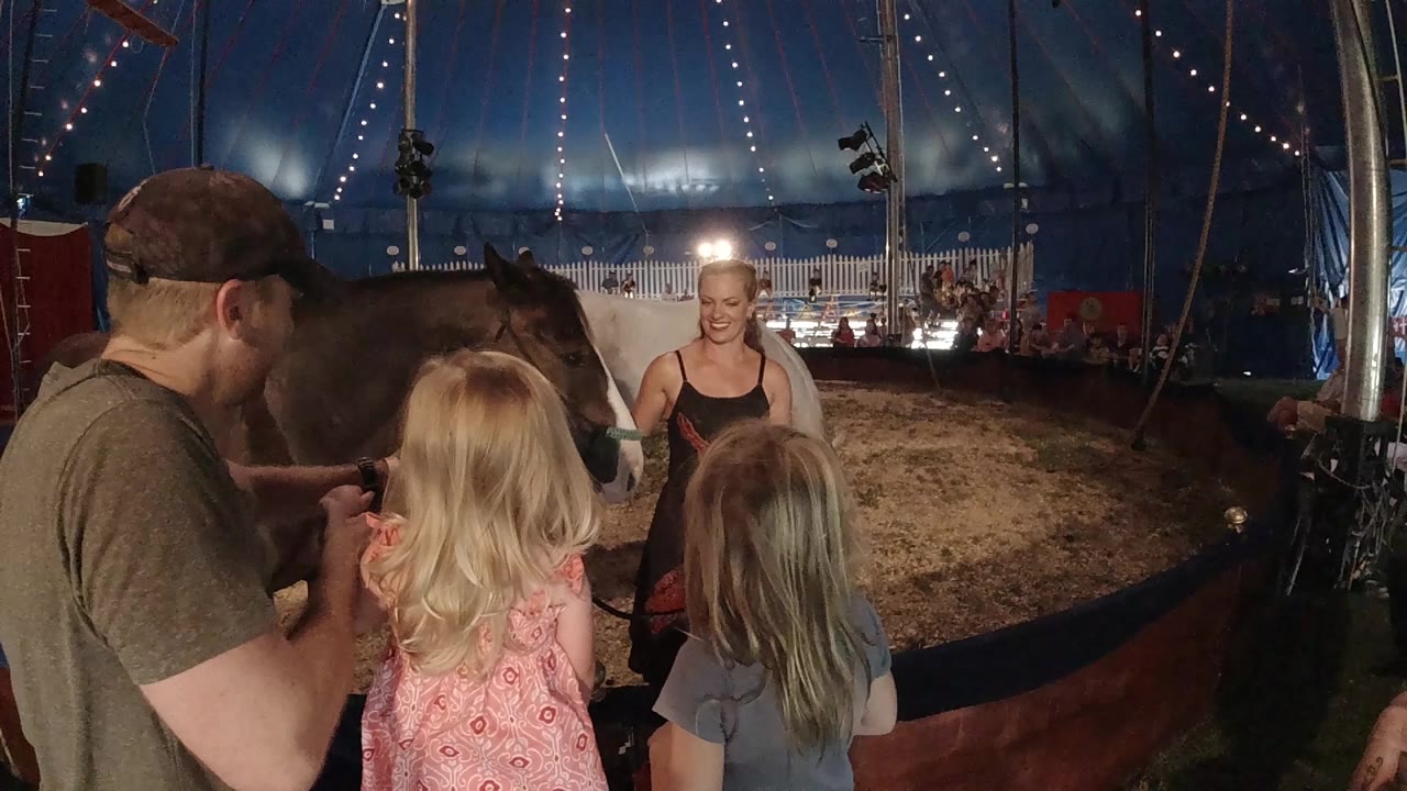 The girls remain in the foreground as the woman stands near the horse, with bright lights and rows of seating visible across the dirt ring.