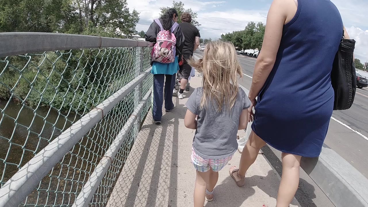 The girl remains near center as the woman walks at the right edge, with the chain-link fence, bridge rail, and leafy trees visible along the left side in Colorado.