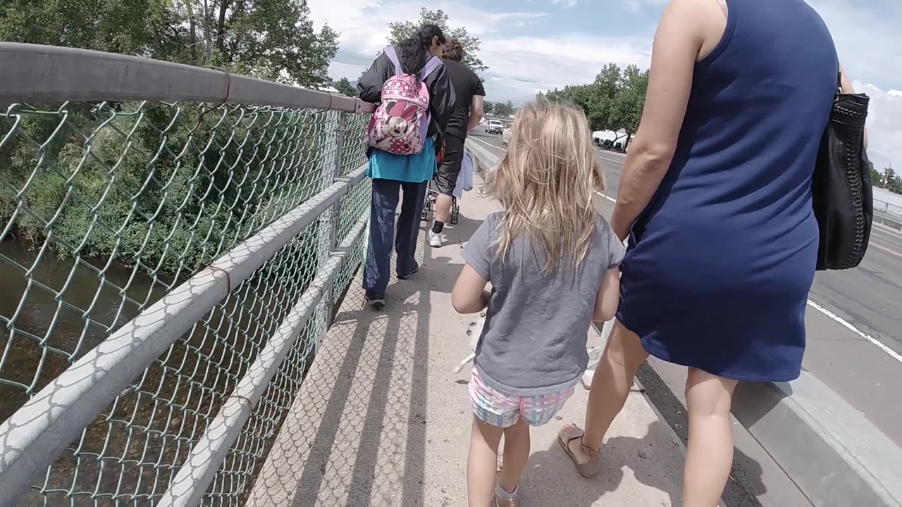 A young girl walks forward between the bridge fence and the woman in blue, while cars and the roadway appear along the right side of the Colorado bridge.