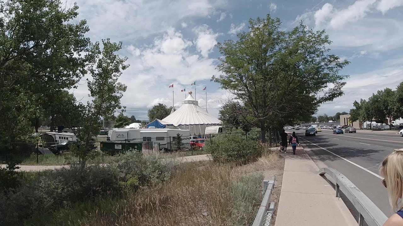 A Colorado roadside view shows the white circus tent behind dry grasses and a fence, with pedestrians walking ahead on the sidewalk.