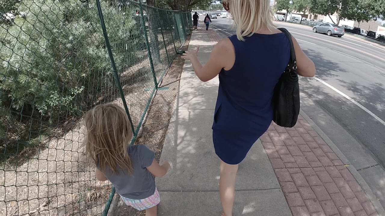 The woman steps forward with her black bag while the young girl keeps close to the green fence on the tree-lined Colorado sidewalk.
