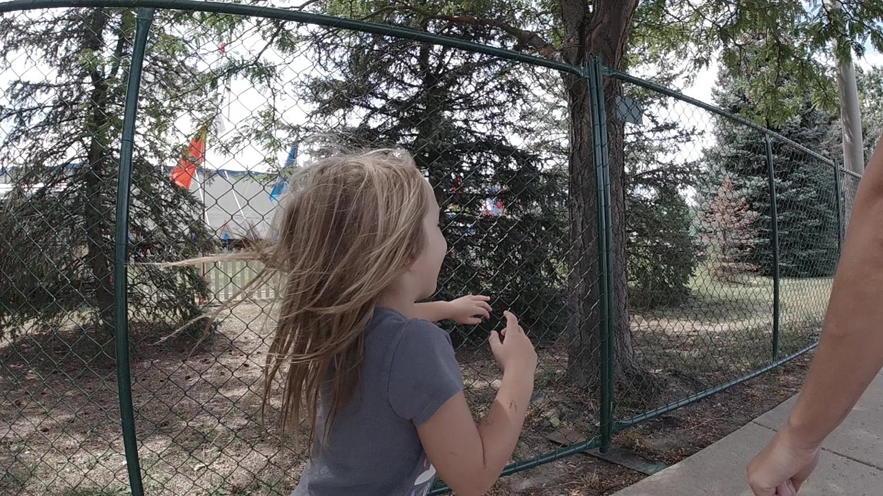 Blonde girl beside a chain-link fence and trees.