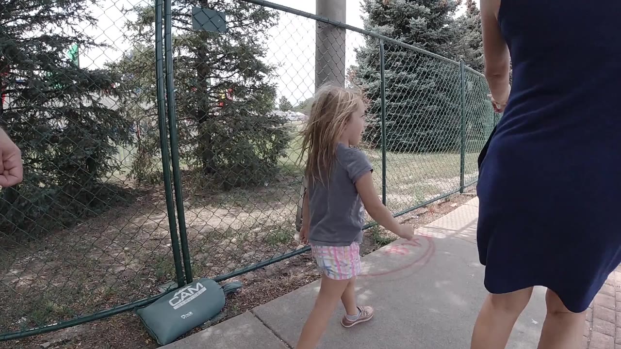 Young girl walking beside a cropped adult near a chain-link fence.
